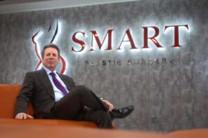 Dr. Jeffrey Weinzweig sitting on an orange couch at the Smart Plastic Surgery clinic, with the clinic's logo on the wall in the background